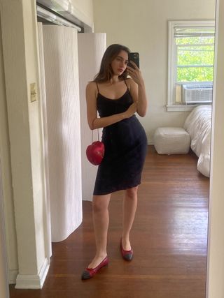 woman taking selfie in mirror wearing black body con slip dress with red heart purse and red black cap toe ballet flats