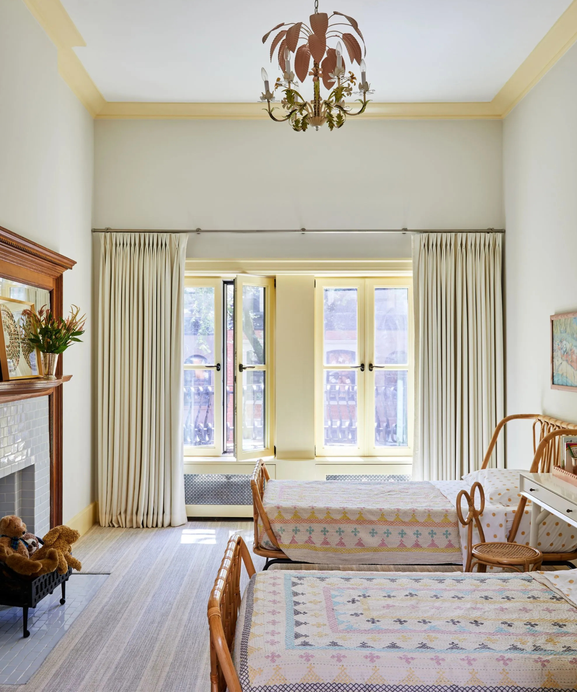 Tall, white bedroom with twin rattan beds, pale yellow window trim, a tiled fireplace with teddy bears, and a delicate wood chandelier