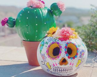 Two pumpkin decorating ideas. In foreground a day of the dead inspired face, in background a trio of pumpkins depicted to be a cacti plant in brown pot