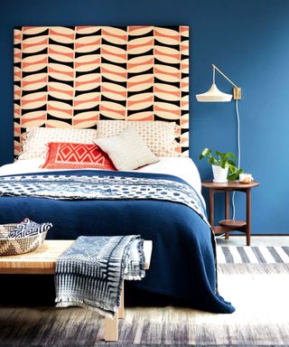 : A bedroom with a tall headboard feature, mid blue painted walls and patterned throws and bedding. A basket and bench seat.