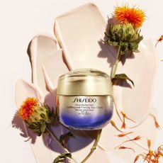 Photo of Shiseido day cream with gold and purple bottle on pale pink background with two single orange flowers