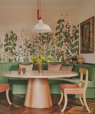 Dining corner with banquette in green velvet and a round wooden table and chairs and a floral tiled mural behind