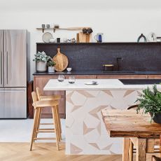 A modern kitchen with American-style refrigerator, island with encaustic tile detail, wooden bar stools and dining table, and black splashback