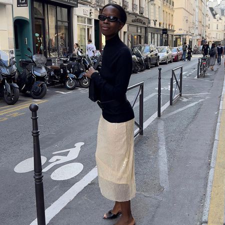 Sylvie wears a black sweater, beige skirt, black heels, and black sunglasses.