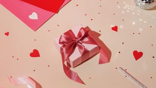 A light pink gift box with blush pink bow, red hearts and confetti on a pastel pink background.