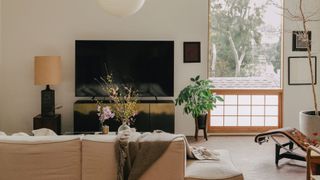 A living room inspired by Japanese interior design, with a potted houseplant, vases of flowers, a tall potted tree, a beige couch, a green throw, a rice paper pendant lamp, and a TV