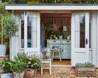 A pale gray National Trust shed demonstrating storage ideas for sheds, with vintage painted furniture and potted plants in front.
