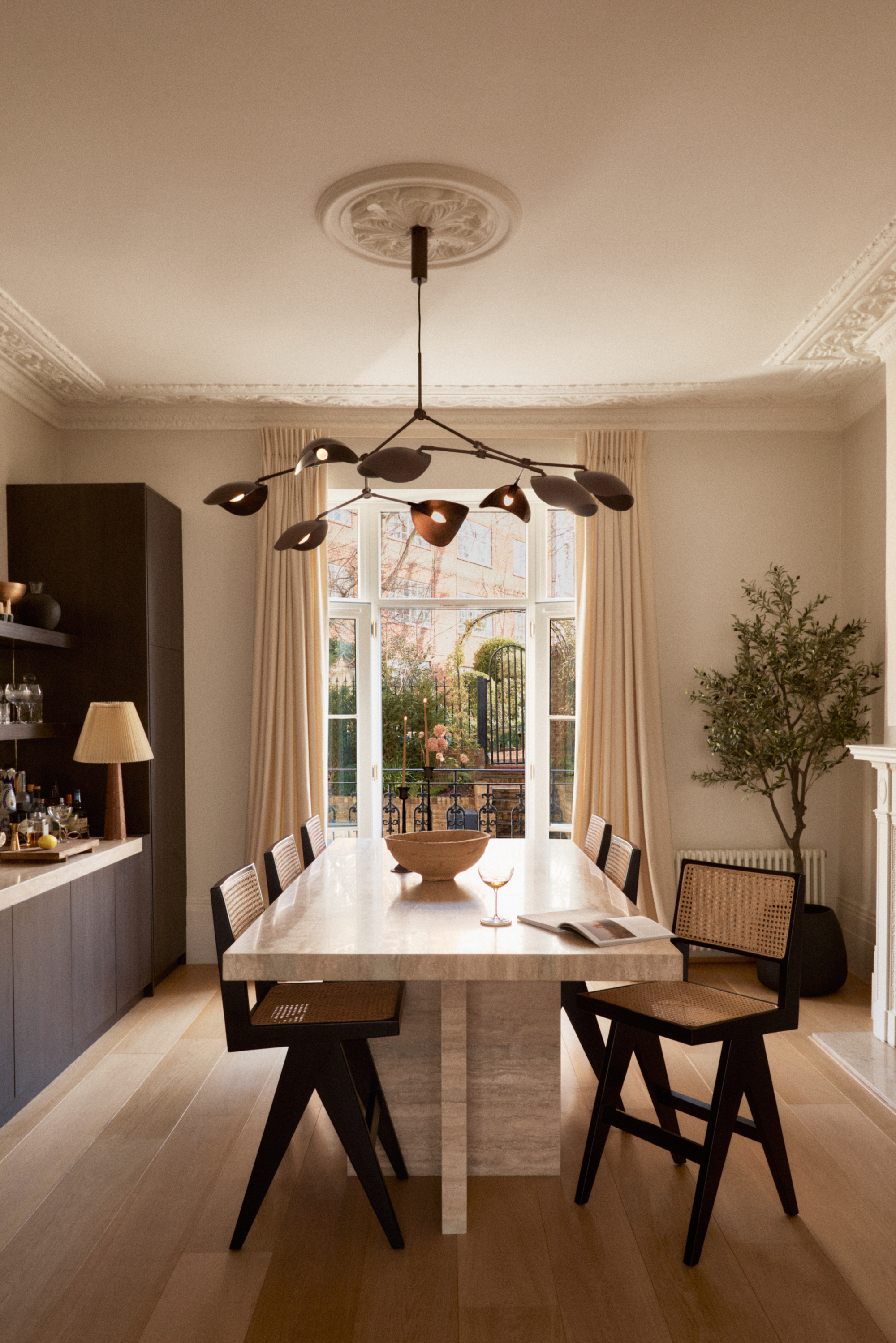 An elegant dining room featuring a large stone dining table surrounded by cane-back chairs, topped by an organic branch-like chandelier in front of large windows opening to a courtyard garden