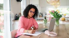A woman works on paperwork and a laptop at her dining room table.