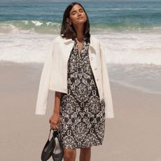 Woman wearing Hobbs black and white dress, holding heels with the beach and sea behind her