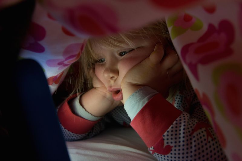 A toddler plays on a tablet underneath a blanket.