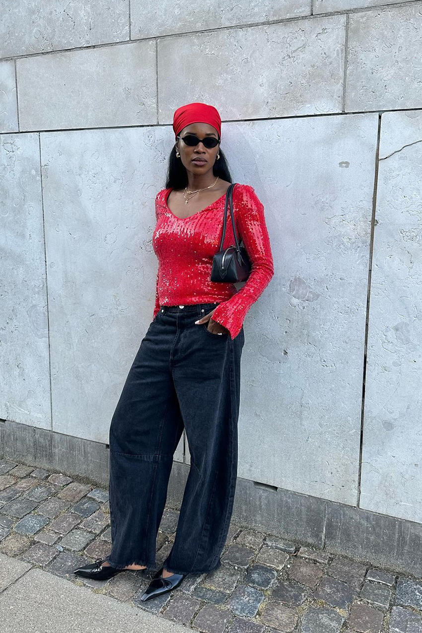 How to wear a bandana is shown in a photo of a woman outside leaning against a concrete wall wearing a red kerchief, black sunglasses, and a red sequin top, a black shouder bag, black baggy wide-leg jeans, and black pointed kitten heels