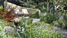 A relaxed seating area is surrounded by flower beds filled with yellow and blue perennial plants at the Chelsea Flower Show