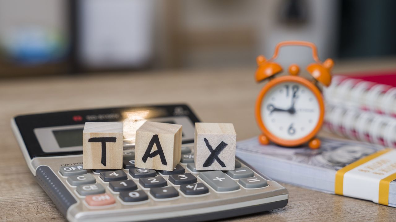 Wooden blocks spell out the word tax on top of a calculator next to an alarm clock sitting on a stack of cash.