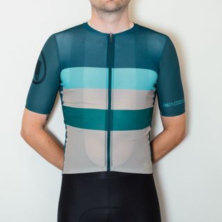 A white man in a striped grey and green cycling jersey and black cycling shorts stands against a white wall