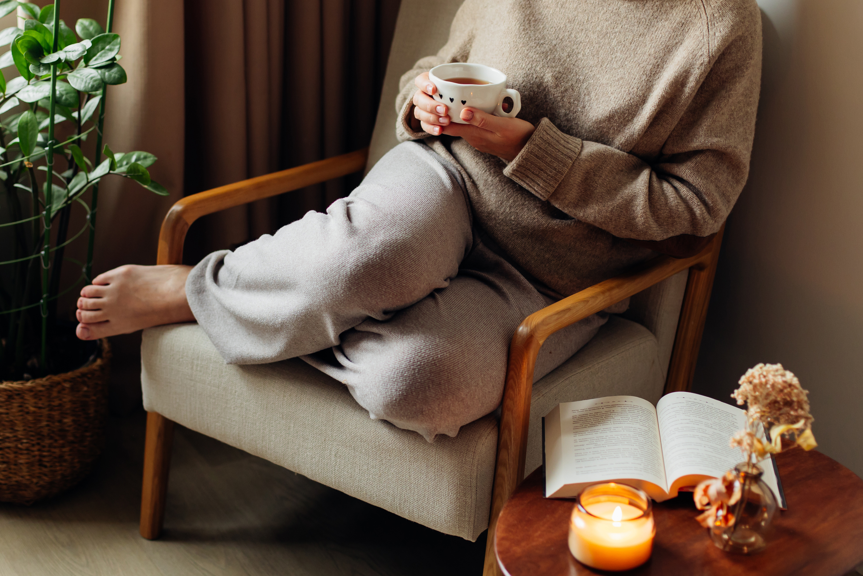 A person wearing loungewear while sitting in a chair alongside a small, wooden side table with a book and a candle.
