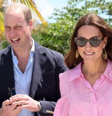 Prince William wearing a blue open necked shirt with a navy blazer standing outside next to Kate Middleton in a pink dress and gold hoop earrings with sunglasses