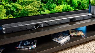 The Polk Audio Signa S4 being used in a lounge with a large TV. The rear of the soundbar is on display showing the ports. On the TV screen is a green forest scene. In the background is a blue wall.