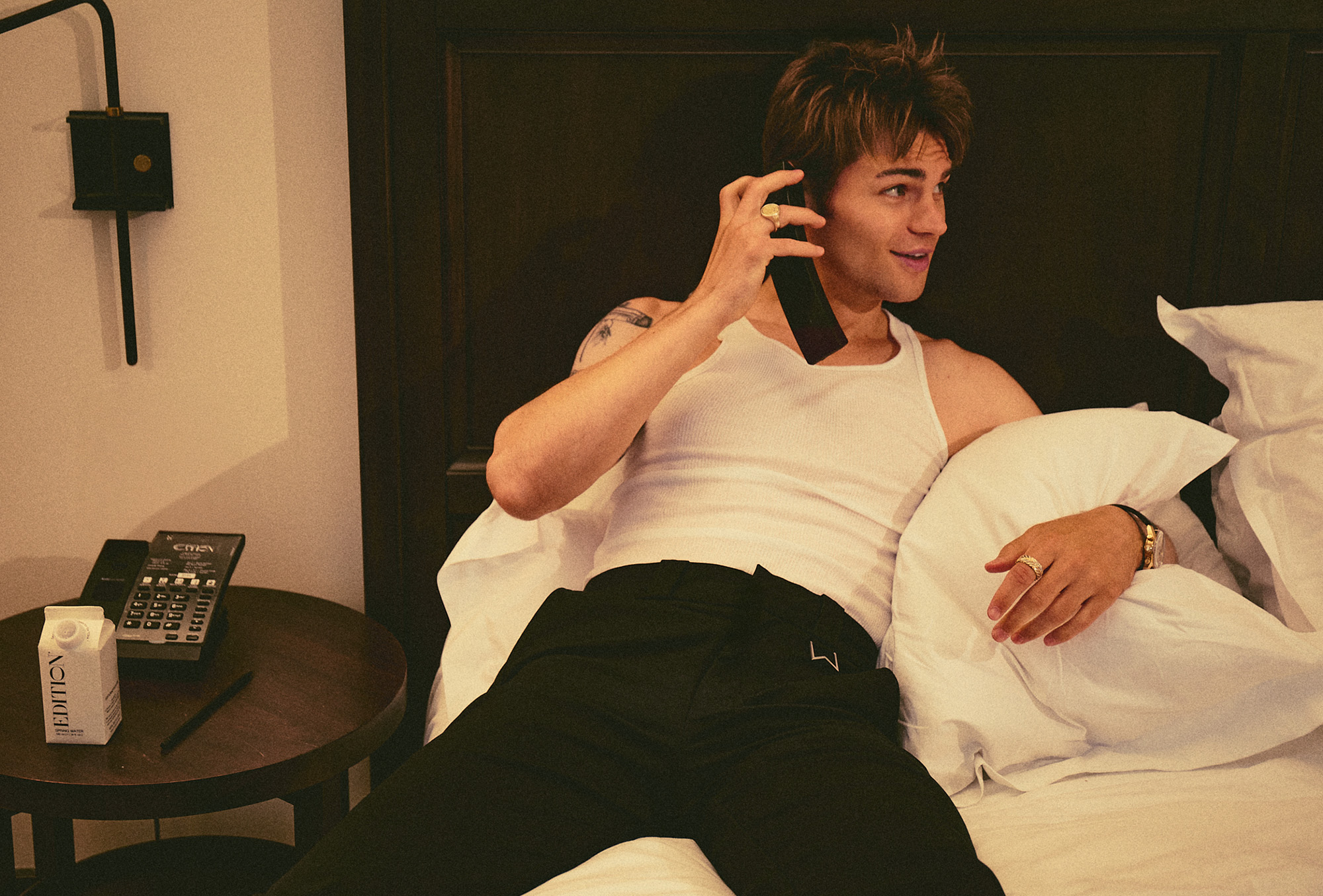 My Life With the Walter Boys actor Noah LaLonde laying on his hotel room bed talking on the phone. He is wearing a white tank top and black pants by Rhude.