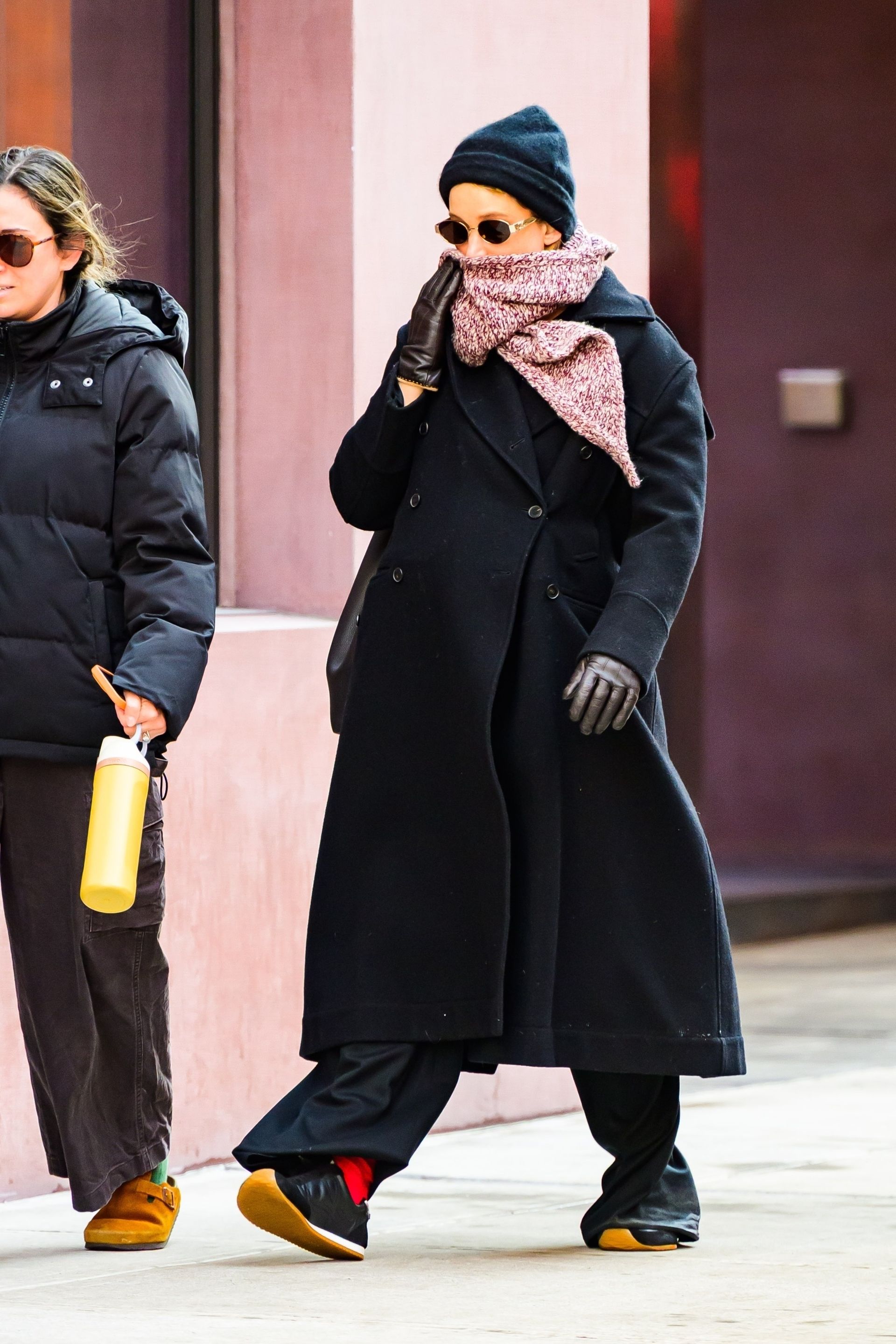 Jennifer Lawrence wears a black beanie, sunglasses, red scarf, black jacket, black Loewe sneakers, and red socks.