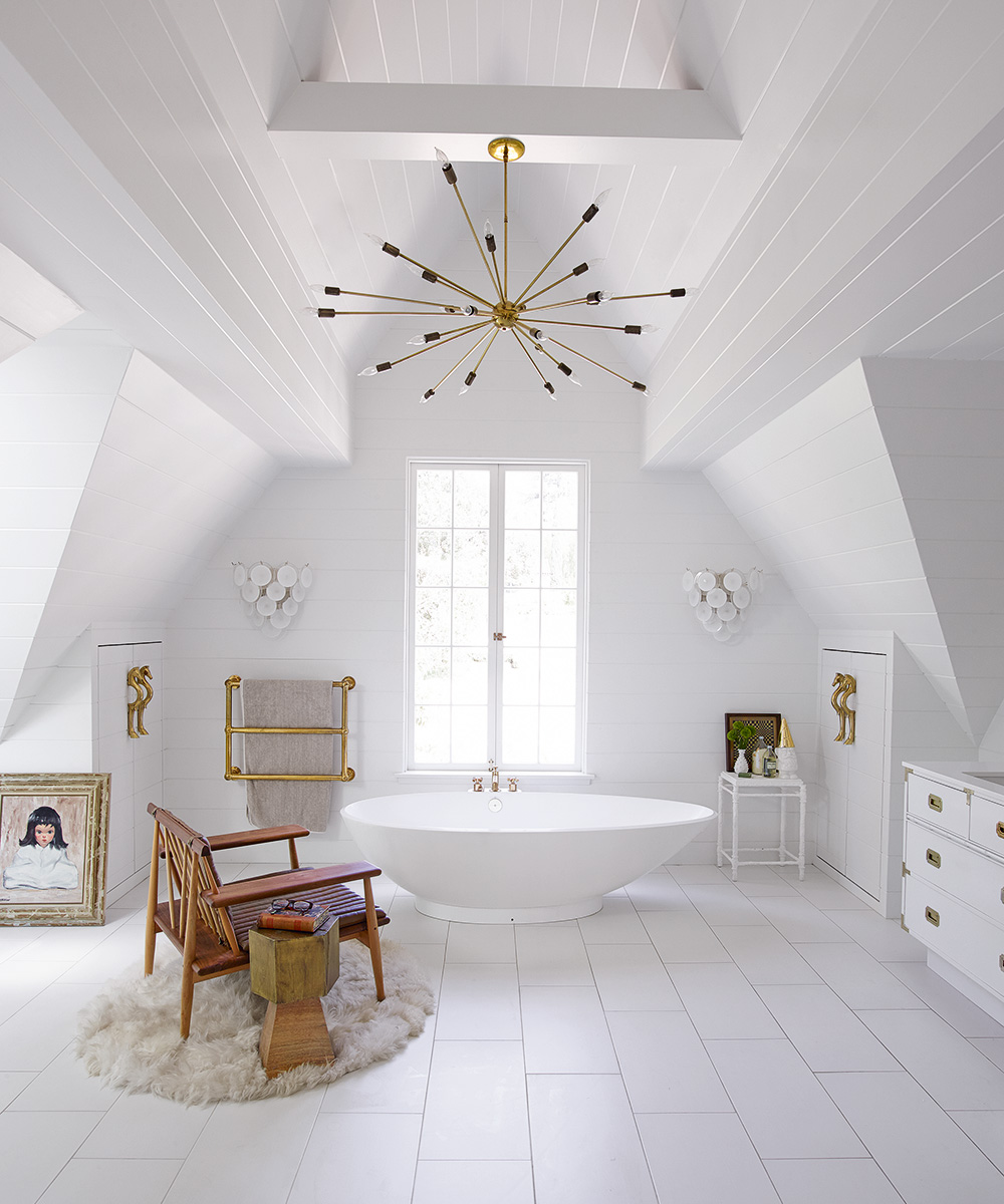 A large with bathroom with a white bath below a window, a wooden armchair and white floor tiles