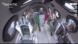 Three people in a space plane. One is holding an Italian flag.