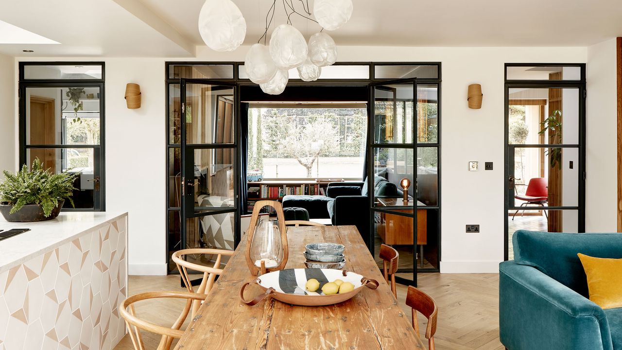 view across wood kitchen table to glass partition and open door to sitting room with steel framed glass door to hall and geometric tiled island and green sofa in view