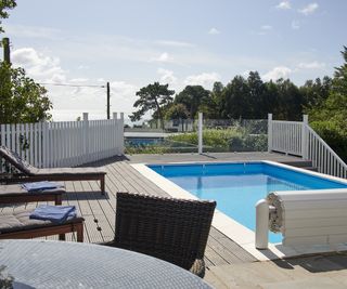 A small pool on a raised deck area surrounded by a white picket pool fence and sun loungers