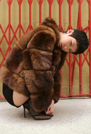 One of the best jacket coat brands, Apparis, is shown in an image of a model wearing a faux fur coat with black shorts and black ankle boots