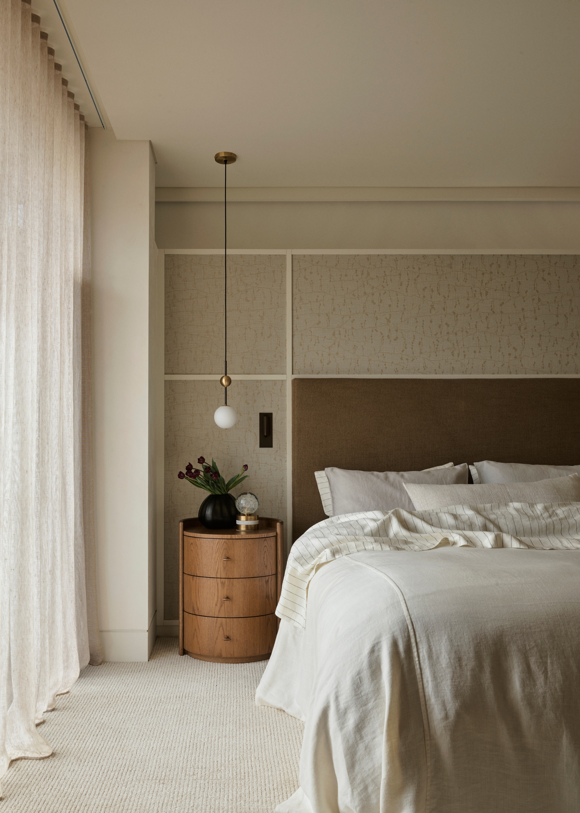 A bedroom with sheer window treatments, a pendant bedside lamp, a curved wood side table, a vase of tulips, and a striped duvet