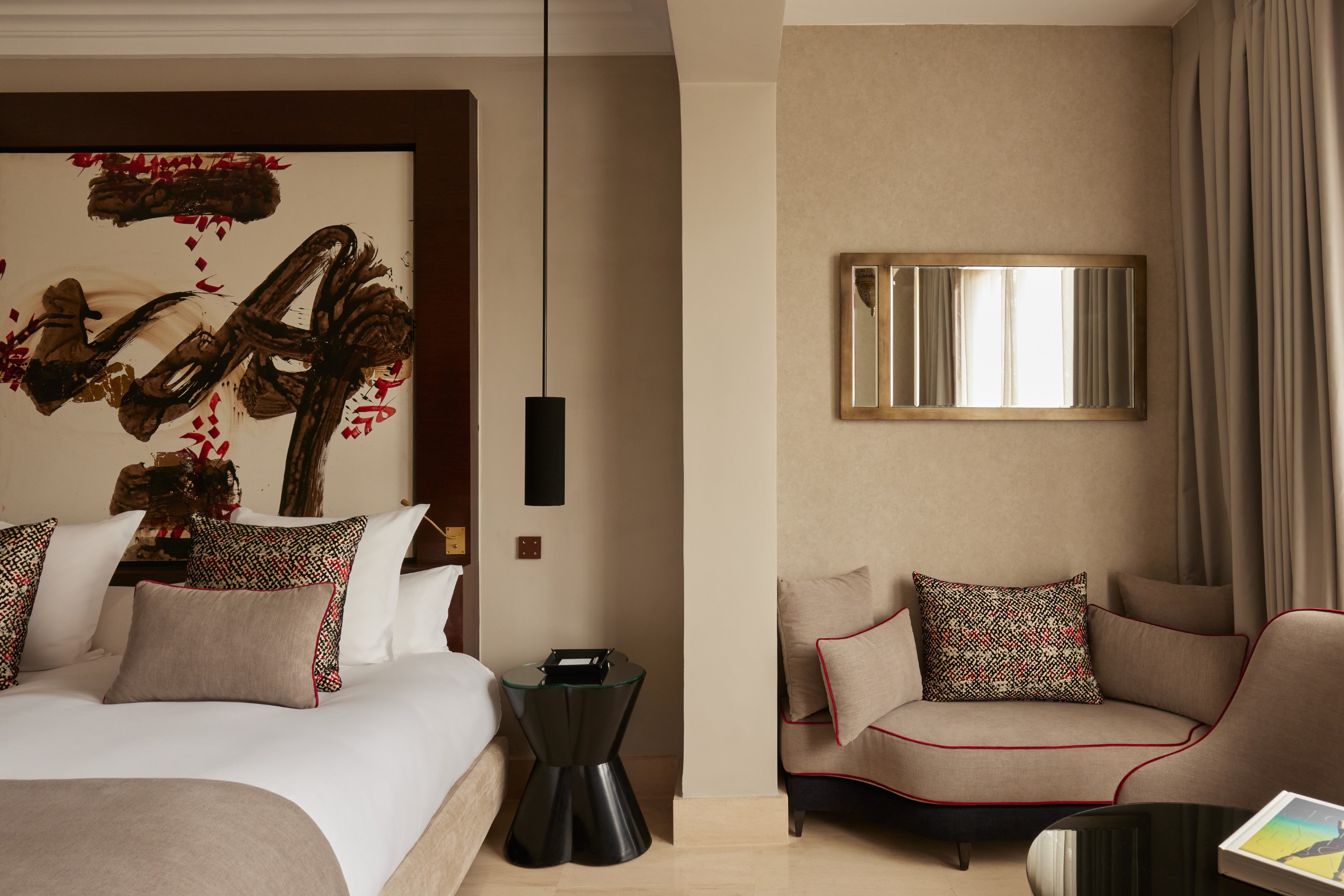 A hotel bedroom with a large abstract artwork as a headboard, soft cushions in pale brown and red, brown, and black patterns, a bedside table sculpted in lacquered black wood, and a mirror.
