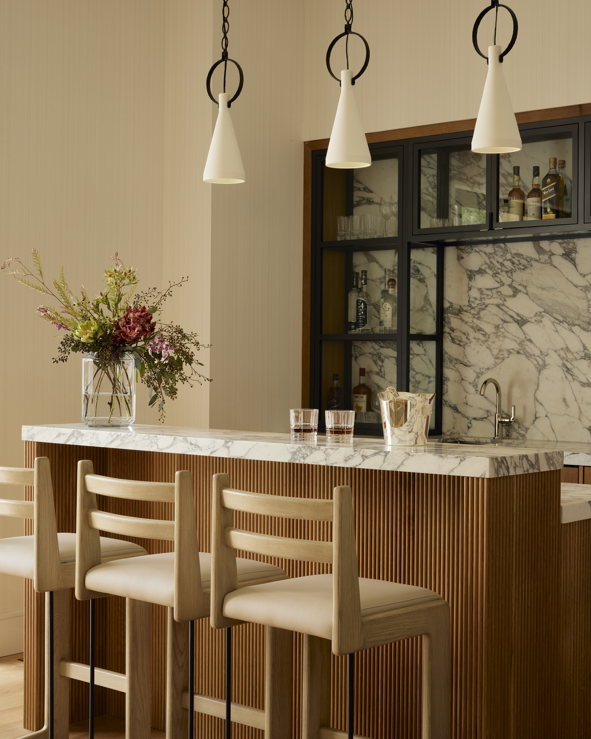 modern kitchen with raised countertop with bar stools, flutted joinery, a marble countertop and backsplash, three pendant lights hanging overhead, a vase with flowers, glass and steel shelving styled with bottles