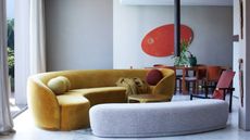 gray living room with curved gray ottoman, curved mustard velvet sofa, red round sculpture on wall, red armchair, round pillows, and mustard yellow throw