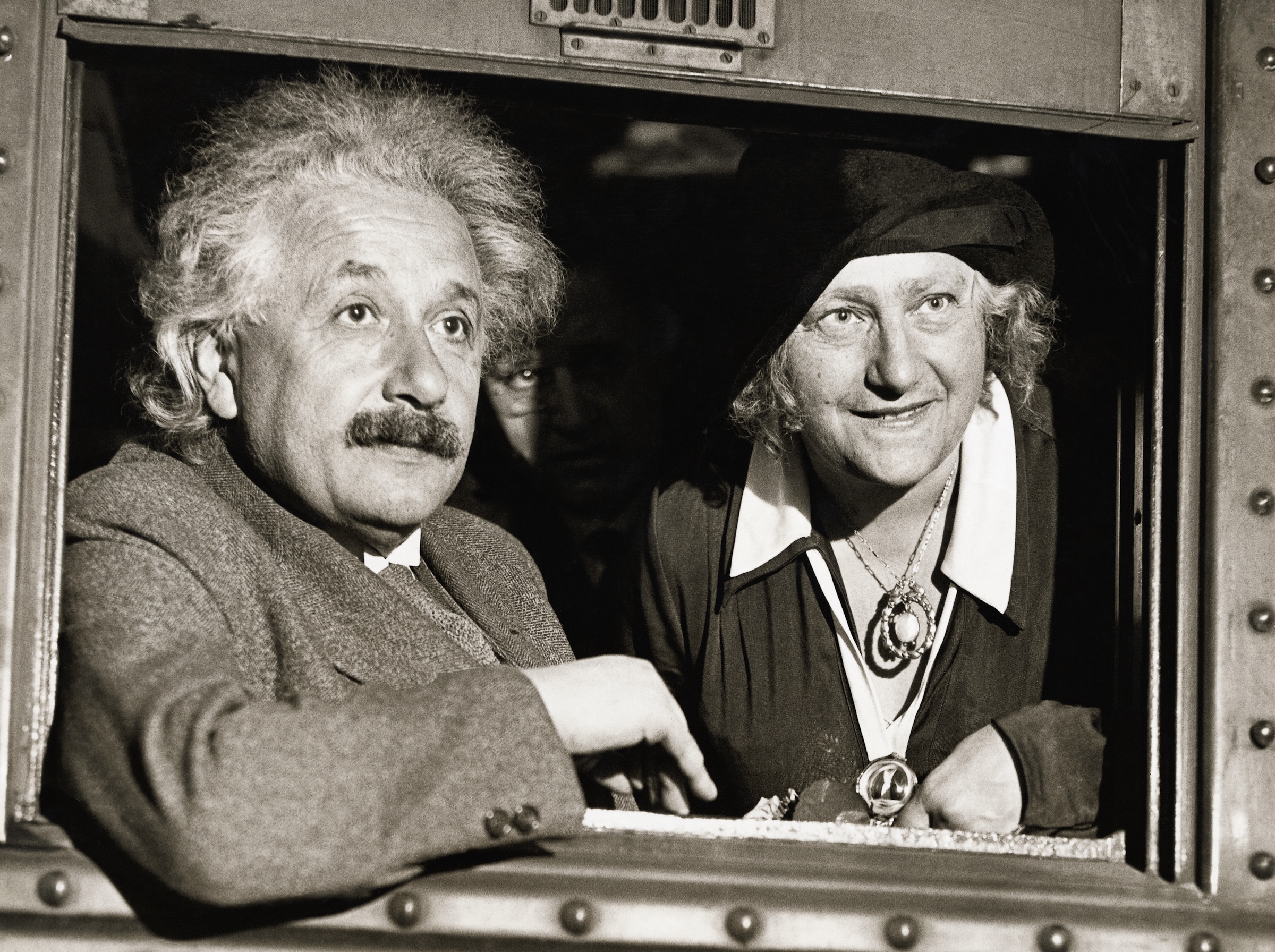 Albert Einstein and his wife look out of a window in a black and white photo.