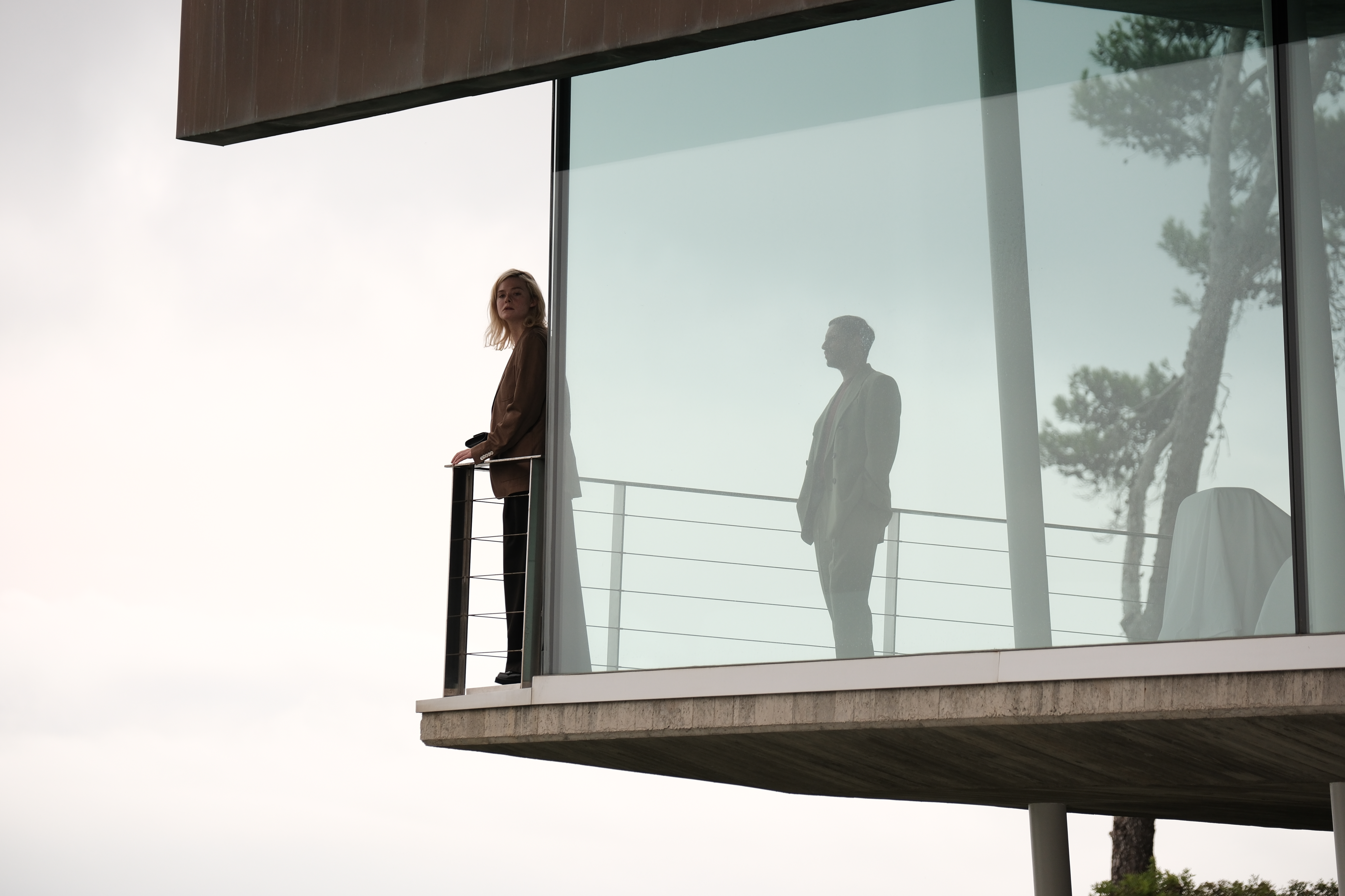 Film still depicting the mid-century modern exterior of a glamorously designed wood, stone, and cement villa, inhabited by a family dressed up in sumptuous clothing and interacting across its rooms.