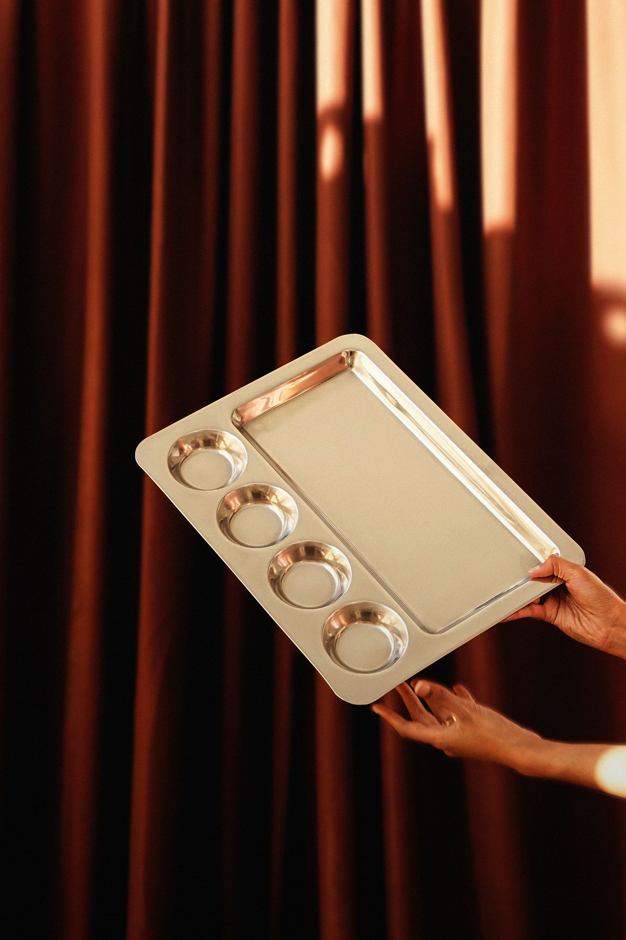 A silver plate is held by two hands in front of a terracotta-hued curtain.