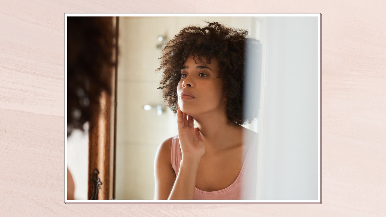 Image of woman touching her jawline while looking in the mirror in a frame against a pink background