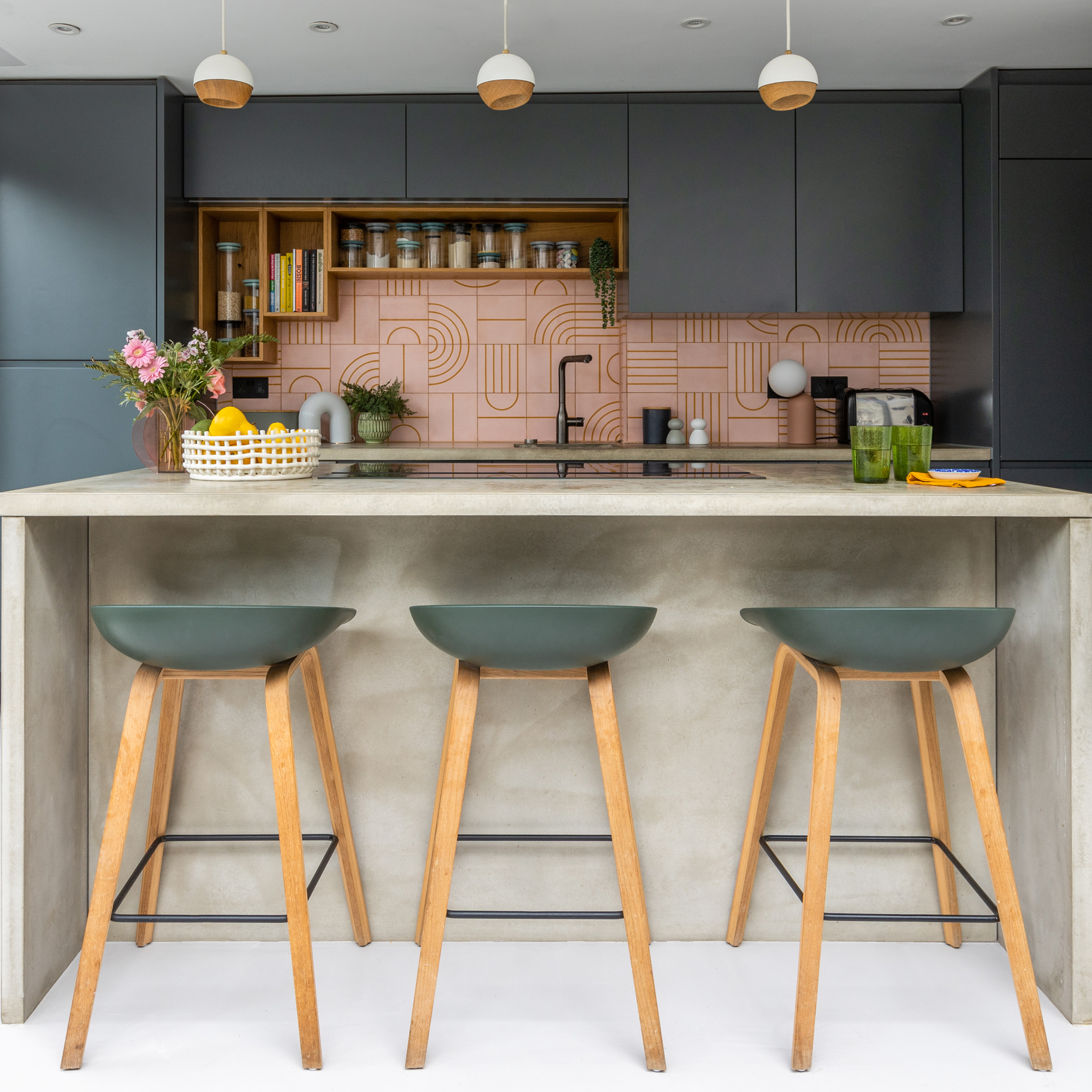 a contemporary dark blue kitchen with concrete kitchen island, bar stools and pendant lights with a pink graphic tiled splashback