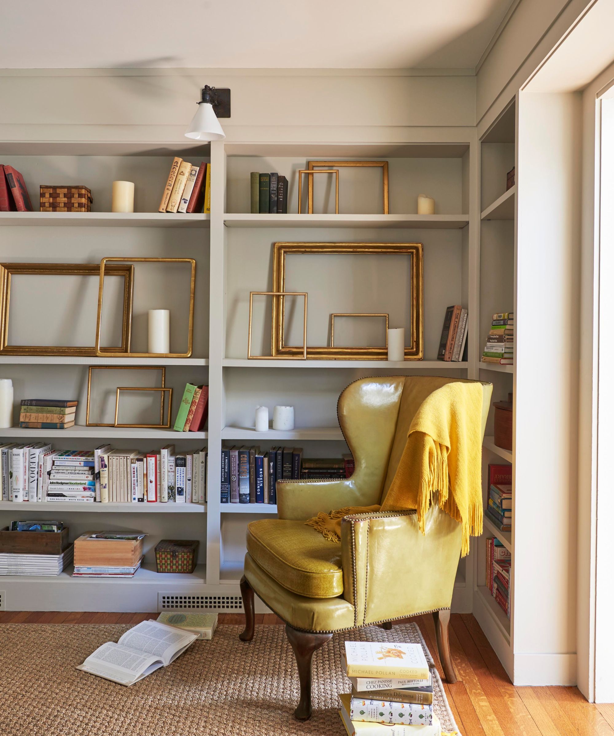 Home library with built-in bookshelves painted in Benjamin Moore's Edgecomb Gray, books and gold picture frames on the shelves, a yellow leather armchair, and a yellow blanket draped over it.