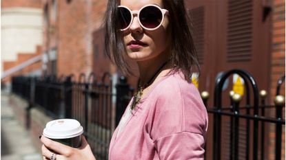 Fashionable young woman with coffee to go wearing pink sunglasses and cardigan - stock photo