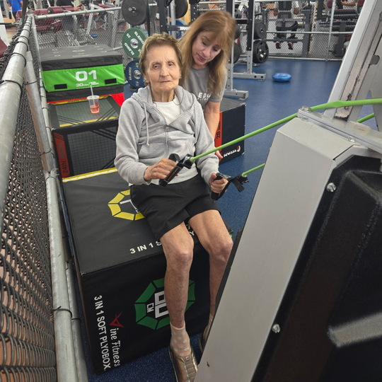 100-year-old Mary Coroneos sits on a gym box and holds a resistance band in her hands, that is looped around a nearby pole. Behind her a personal trainer is adjusting her posture.