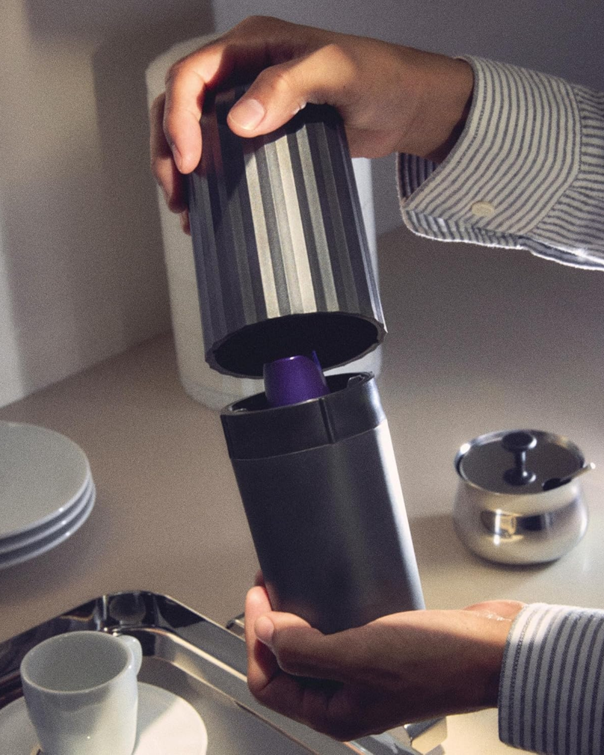 Image of someone's hands holding a black, ribbed coffee capsule crusher in front of a beige countertop. There is a stack of small white plates and a silver dish in view. The image is grainy like film, and there are shadows from the items on the countertop.