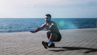 Fit young man doing pistol squats on seafront. Fitness training in morning outdoors. Workout during lockdown outside the gym.