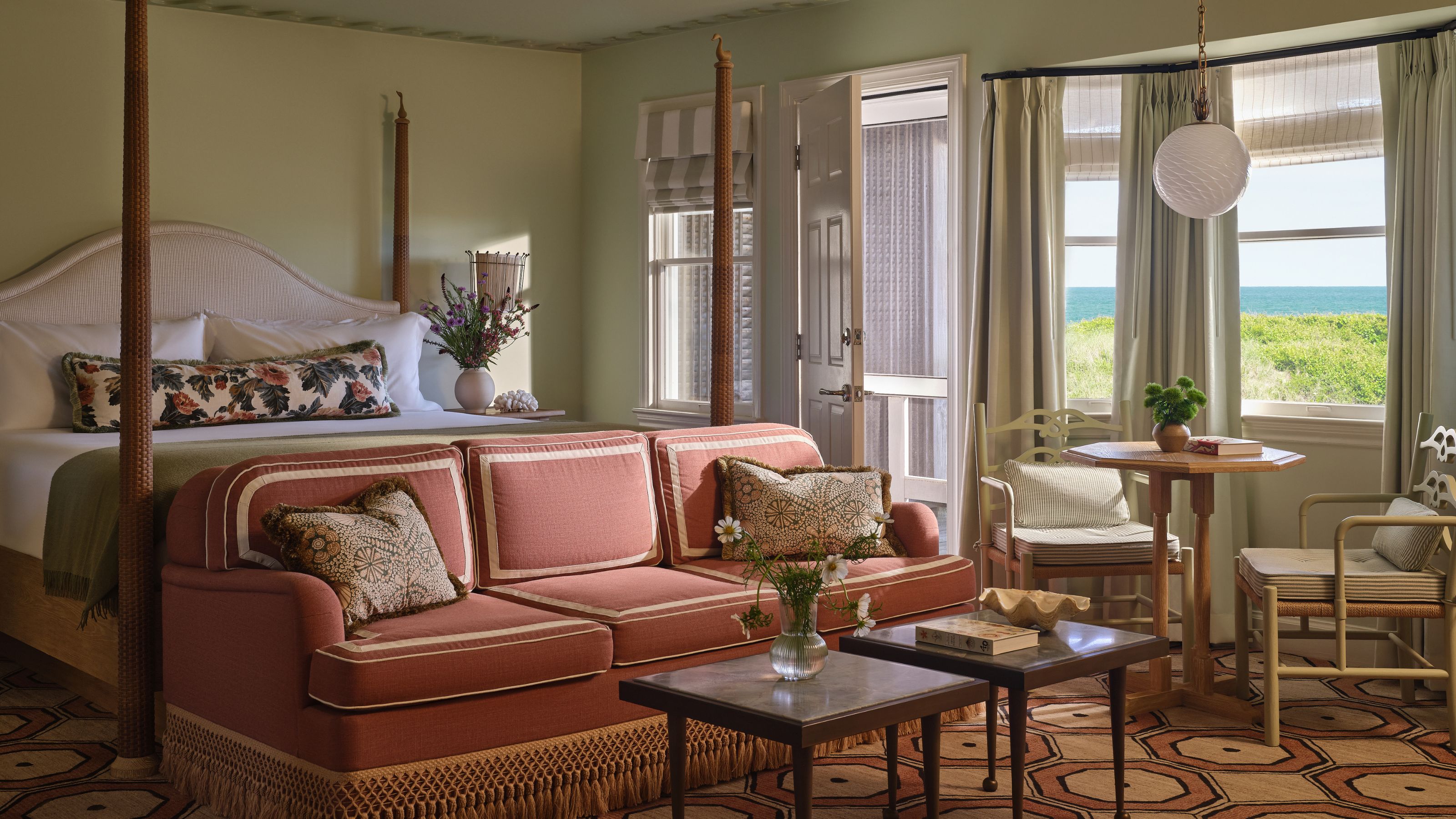 A coastal bedroom with a four-poster bed, a dusty pink sofa at the foot of the bed, and a large window offering a scenic ocean view.