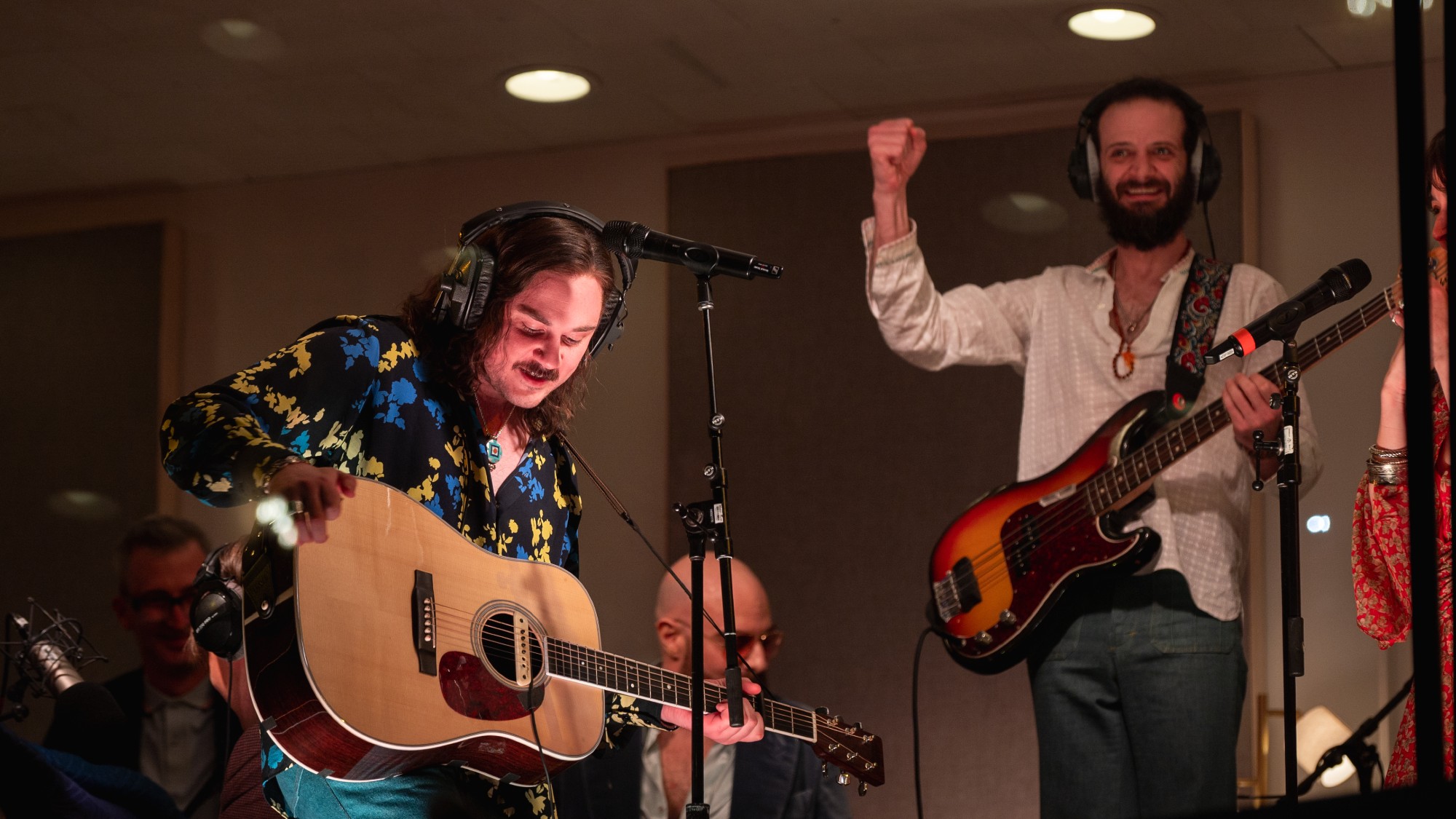 two men play guitar and bass in front of microphones. they are dressed in clothes from the 1970s.