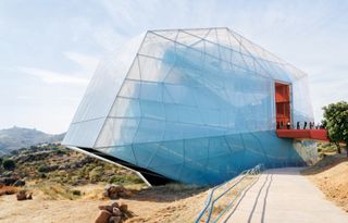 Plasencia Auditorium and Congress Center, blue cloudy sky, surrounding rocky terrain, neutral colour pathway, red bridge leading into the building with visitors walking along, tree to the right