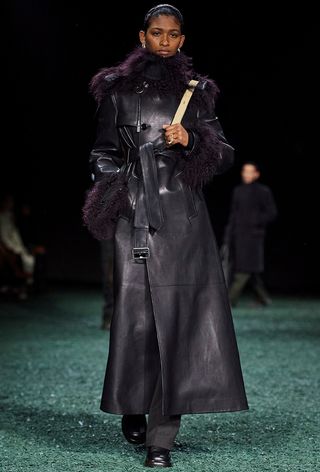 Model walking the Burberry runway show wearing a leather purple trench with fur, gray trousers, and loafers.