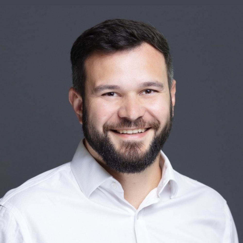 A studio-style headshot of a man with dark hair and a full beard, smiling slightly, wearing a white shirt against a plain dark background.