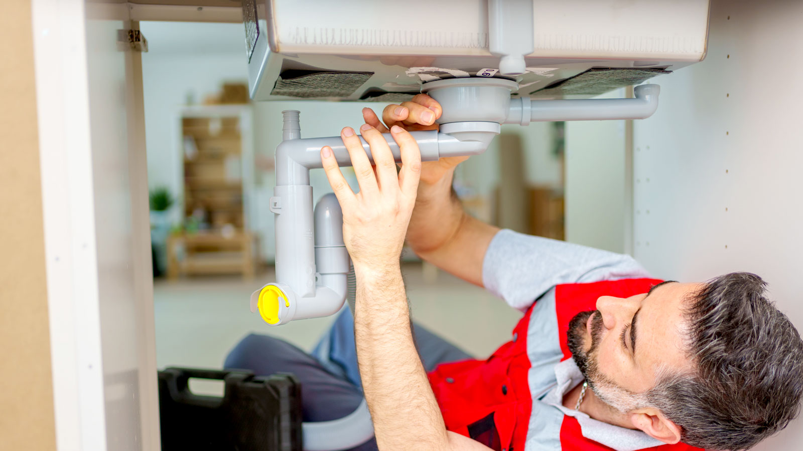 Plumber in red jacket lying under sink fitting a grey kitchen waste unit