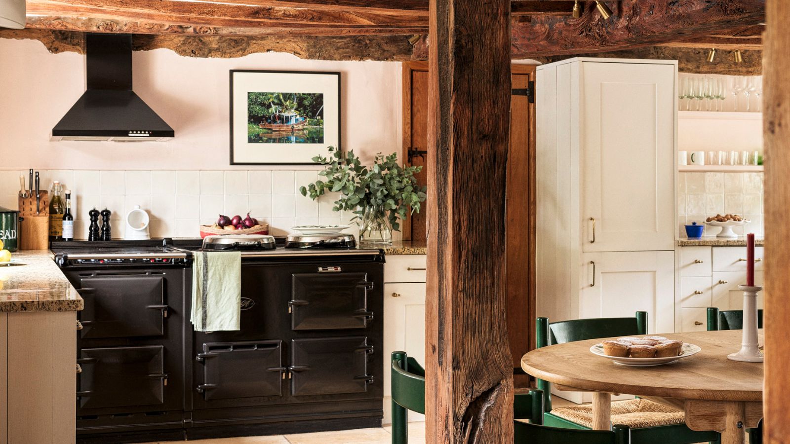 A cozy cottage kitchen with wooden ceiling beams, pink walls, white cabinets, a black AGA, and subtle Christmas decor